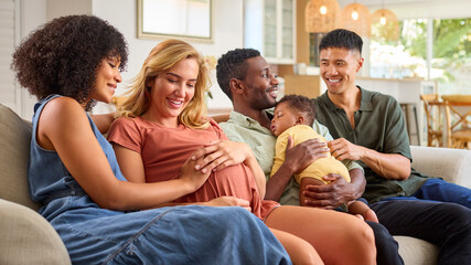 Pregnant Same Sex Couple Visiting Male Couple With Baby Daughter Cuddling Her On Sofa At Home