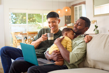 Same Sex Male Couple With Sleeping Baby Daughter On Sofa At Home Working On Laptop