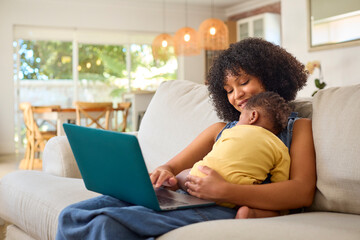 Family With Mother Working On Laptop Cuddling Sleeping Baby Daughter On Sofa At Home