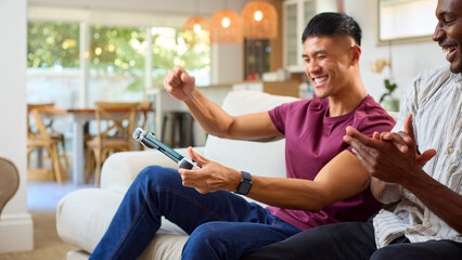 Same Sex Male Couple Or Friends On Sofa At Home With Handheld Mobile Computer Gaming Console