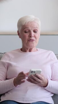 Senior Caucasian happy woman mobile using phone for texting in social media at home. Resting online and spending leisure time. Vertical video.