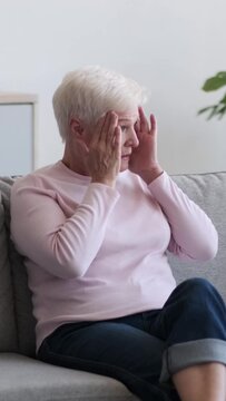 Unhealthy Caucasian senior adult woman with headache, sitting on sofa at home. Illness, annoyance, exhaustion concept. Vertical video.