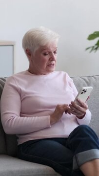 Upset Caucasian old woman using mobile phone for browsing content in internet and reacting on bad news at home living room. Vertical video.