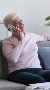 Old cheerful Caucasian woman talking mobile phone and laughing at home. Digital conversation during weekend leisure time. Vertical video.