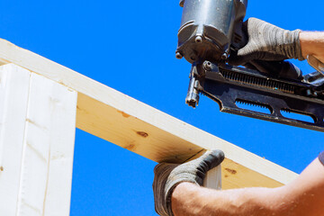 Worker in holding plank installing it on household construction using air hammer in nailing wooden beams.