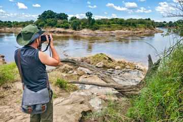 A male photographer in a green hat takes a photo the savannah on the background of river in masai...