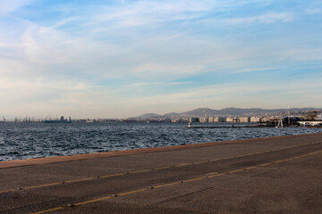 A scenic coastal pathway in Thessaloniki, Greece offers expansive views of the glistening sea with a city skyline and mountains in the distance.
