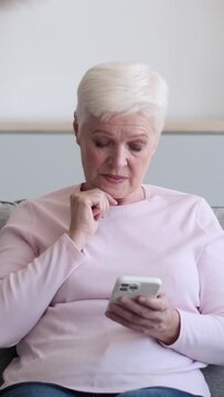 Friendly Caucasian elderly woman using smartphone browsing internet and resting sofa at home. Spending free with online entertainment. Vertical video.