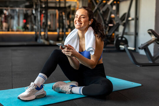 Sports lifestyle. Lady in sportswear uses cellphone during workout, resting and smiling at camera. She posts photos from the training on social media