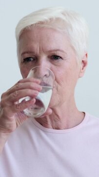 Mature happy Caucasian woman drinking clear water from glass and looking and smiling at camera on grey background. Recreation, recovery concept. Vertical video.