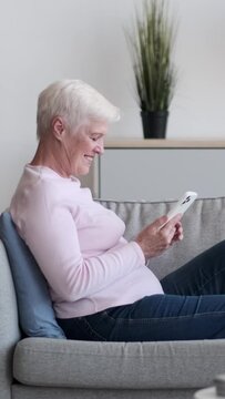 Friendly Caucasian elderly woman using phone, messaging in social media or browsing internet, lying on sofa at home. Leisure time relaxation. Vertical video.