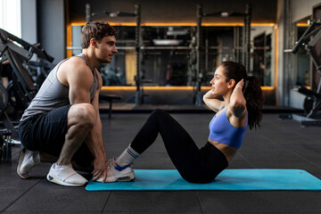 Handsome man trainer assisting young beautiful woman while lady doing sits ups in gym on fitness mat, side view