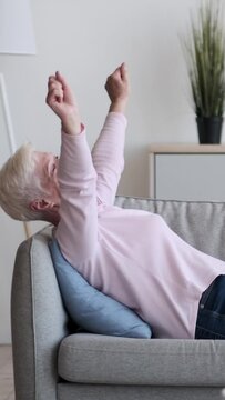 Joyful and excited mature Caucasian woman resting and stretching on couch at home living room. Refreshment, satisfaction concept. Vertical video.