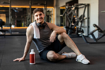 Fototapeta na wymiar Taking break and drinking water. Sportive strong man have workout day in gym, sitting on floor and smiling at camera