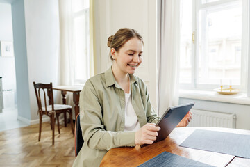 Smiling woman with braces comfortably using a tablet at a dining table in a well-lit, spacious room.
