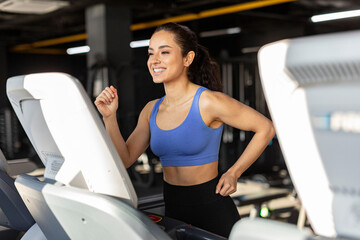 Excited young lady in sportswear exercising on treadmill at gym, active woman enjoying cardio...