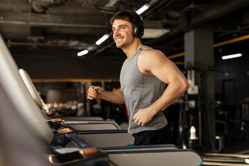 Handsome man in wireless headphones jogging on treadmill at gym and listening music while running, enjoying workout, copy space