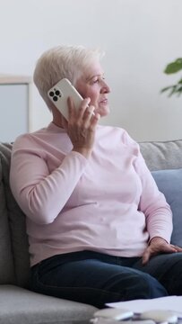 Joyful elderly Caucasian woman talking during mobile phone call at home living room, resting on sofa. Digital communication and contact. Vertical video.
