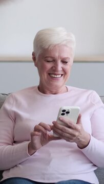 Cheerful senior Caucasian woman messaging in online social media relaxing on sofa at home living room. Spending free time in cyberspace. Vertical video.