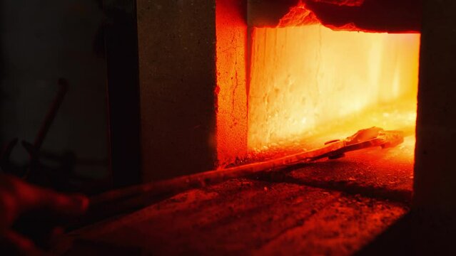 A close up of a stone furnace in a forge, with a red flame flaring up. A metal product is being pushed inside to be heated and melted.