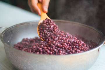 Woman's hand release temperature by using wooden ladle scoop boiled azuki beans in stainless steel sieve before mixed with tempeh starter, isolated on white table, closed up.