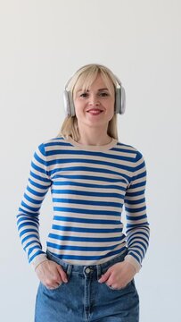 Positive portrait of a cheerful and friendly Caucasian woman listening music using headphones and dancing on white background. Vertical video.
