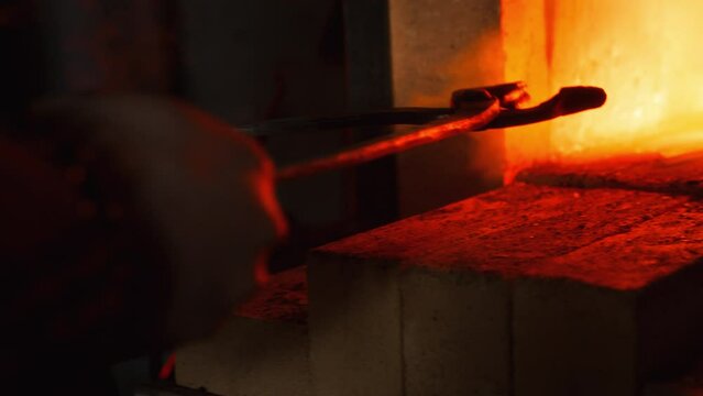 A close up of a stone furnace in a forge, with a red flame flaring up. A metal product is being pushed inside to be heated and melted.