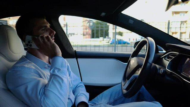 Male entrepreneur speaking by smartphone during riding on electrical auto with autopilot at urban road. Confident businessman talking on phone while riding an autonomous self driving electric car