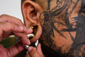 An asian man attaches a large captive bead ring to his flesh tunnel. Example of extensive facial tattoos and body piercings or modifications. - obrazy, fototapety, plakaty