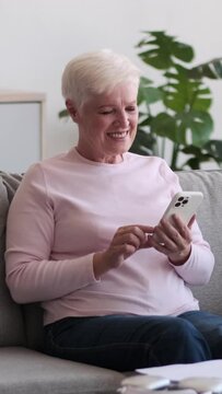 Joyful elderly Caucasian woman with mobile phone resting on sofa at home living room. Online chatting and communication. Retirement and enjoyment concept. Vertical video.