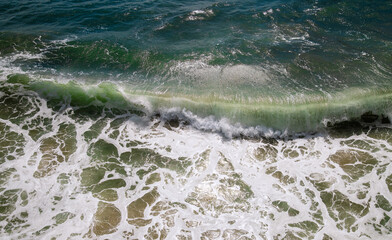 Aerial view of a breaking wave at Santa Monica beach in California, USA. Colorful surf backlit by sun in shades of green, blue and turquoise, with white foam and bubbles. Currents and water in motion.