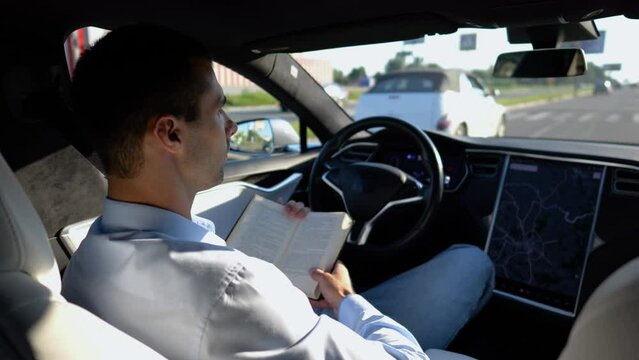 Male businessperson reading book during riding on electrical vehicle with autopilot at urban road. Successful businessman improving his knowledge while riding an autonomous self driving electric car