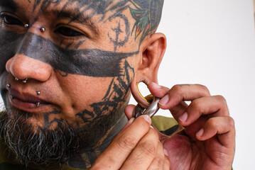 An asian man attaches a large captive bead ring to his flesh tunnel. Example of extensive facial tattoos and body piercings or modifications.