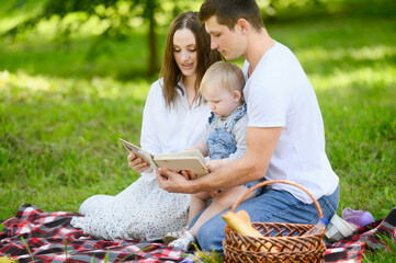 Happy family enjoying a picnic in park and reading a book to their baby. They are all smiling,...