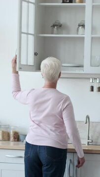 Elderly happy Caucasian woman setting dinner table at home kitchen. Serving with tableware and preparing to banquet. Vertical video.