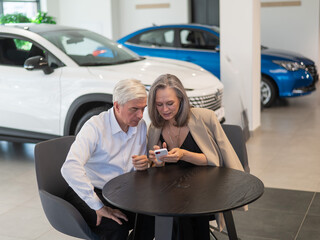 A mature couple is sitting in a car dealership and looking at a smartphone. 