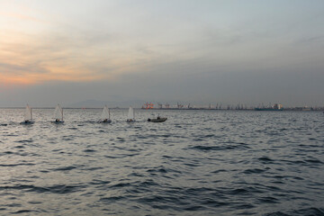 Choppy sea waters under a twilight sky with silhouettes of boats and a distant, faint city skyline...