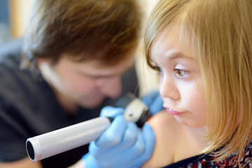 A caring doctor checks moles on the skin of a small child. A dermatologist looks at a rash on the back of a girl using a dermatoscope.