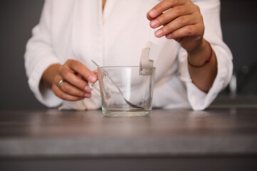 Close-up woman's hands holding a tea bag over an empty glass cup in the home kitchen, preparing hot...
