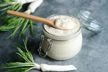 Handmade horseradish sauce in a jar with a spoon on top, horseradish roots on a gray background....