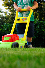 Lower body of a boy in shorts pushing a colorful plastic children's lawn mower on a green lawn....