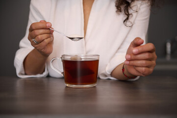 Close-up woman's hands holding a teaspoon with white sugar above a glass cup of freshly made black tea in the home kitchen, preparing hot drink for breakfast in the morning