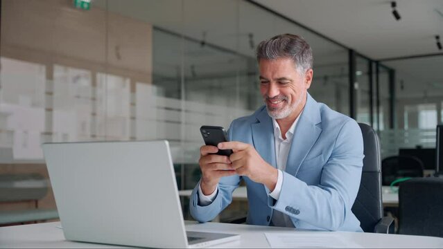Older businessman professional entrepreneur looking at smartphone at work making baking payment. Happy busy middle aged business man ceo investor wearing suit sitting in office using mobile cell phone
