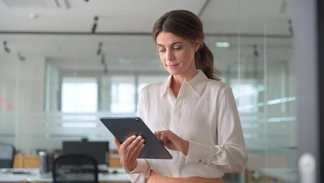 Busy middle aged mature professional business woman banking manager, 45 years old female executive or entrepreneur holding tab digital tablet standing in office at work, looking at pad.