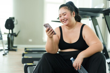 Chubby Asian woman exercise in gym, smiles while talking on phone, taking joyful break. With a...