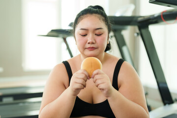 Chubby Asian woman exercise in gym, female contemplates her snack, reflecting on her dietary...