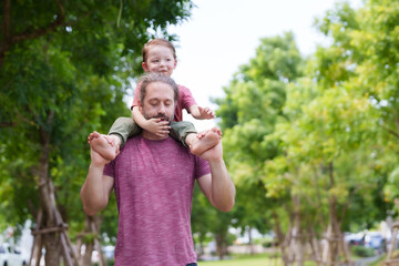 Obraz premium Man carries child on shoulders, walking in park, trees in background, child plays with father's hair, both content. relaxing day for father and son on the weekend.