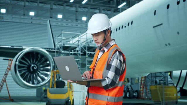 Side View Of Asian Male Engineer With Safety Helmet Working On A Laptop While Standing With Aircraft In The Hangar