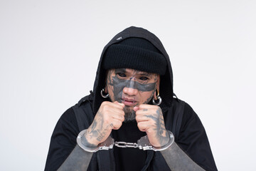 An Asian man with densely inked face tattoos, looking sideways in handcuffs, against a plain white...