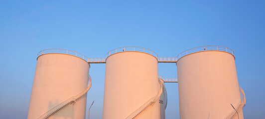 Row of storage fuel tanks with orange sunlight on surface in oil industrial area against blue evening sky background, low angle and panoramic view